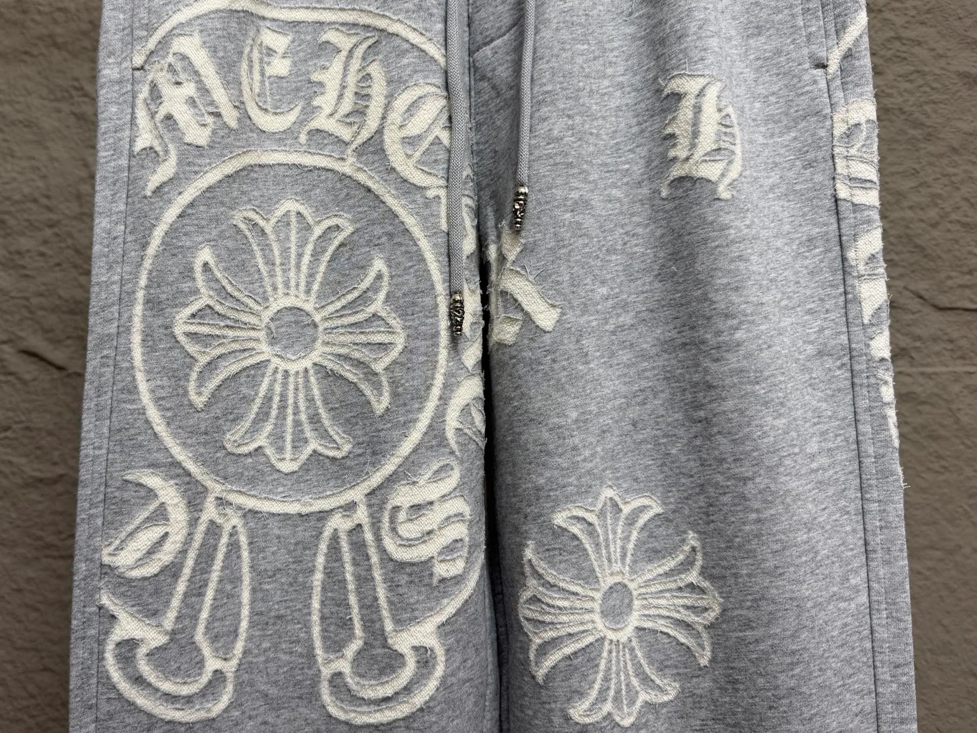 CH/Cro Heart 2026ss Burnt Flower Straight-leg Sweatpants Classic heavy-weight sweatpants with gray tones, Retro street style full-fitting, direct recognition, same style for men and women. ●Three-dimensional patchwork technology: The whole body is covered with Gothic letters + cross flower totems, using raw edge patchwork embroidery, with a mustache-like old texture, and uneven and layered patterns. It is not an ordinary print, and it is durable and not easy to fall off. ●Style tailoring: Loose, straight and wide-legged version, modifying the shape of the legs, hiding the flesh neatly, not flattering the figure, with an online drape, and the upper body has a relaxed street tone. ●Fabric and waist: Heavy knitted terry fabric, thick and soft, with good drape; elastic drawstring waist, with branded metal engraved rope ends, easy to put on and take off, and strong waist adaptability. ●Detailed design: The side pockets are convenient for carrying items, and the back pocket is decorated with cross-pattern patches. The front and rear patterns echo each other. Every detail is highly recognizable to the brand. ●Wearing scenarios: Suitable for street wear, daily outings, and trendy wear. Paired with sweaters, denim jackets, and leather jackets, you can easily create an American retro cool style. ●The overall gray base is paired with Cream White patches, which are low-key yet impactful. They are both comfortable and trendy. Versatile sweatpants, a must-have for fashionistas, are also available at the counter. SIZE：M L XL XXL XXXL